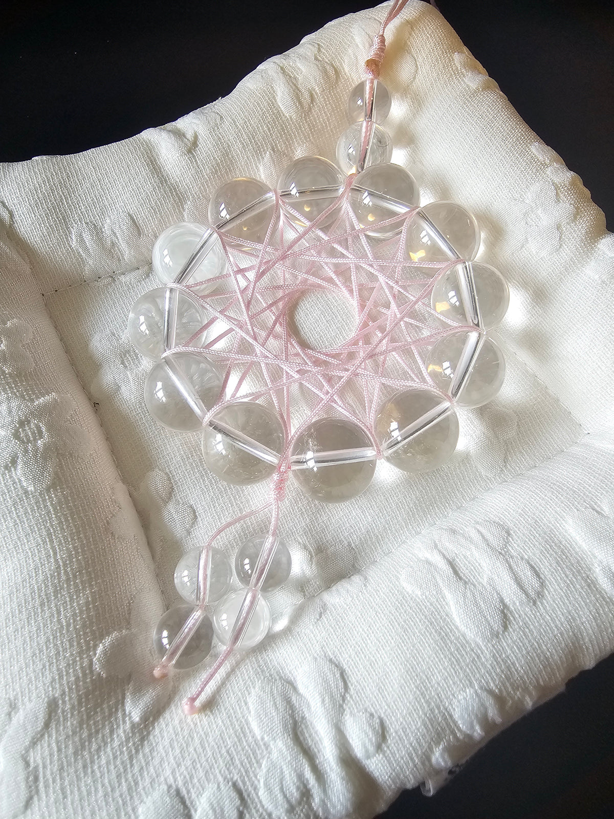 A clear crystal dreamcatcher with a pink woven center and hanging beads, displayed on a textured white fabric surface.